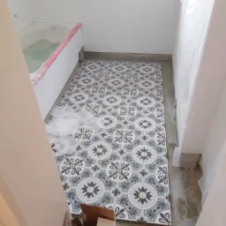 Układanie wzorzystych płytek podłogowych w łazience, widoczna wanna zabezpieczona folią, pojemnik z zaprawą i kartonowe pudło.