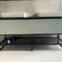 Łazienkowa szafka pod umywalkę z dwoma prostokątnymi umywalkami nablatowymi, fronty szafki w odcieniach szarości z pionowymi panelami, czarna metalowa konstrukcja z półką z siatki, na której leży...