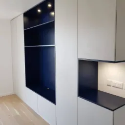 Biała szafa wnękowa z granatowymi wnękami, oświetleniem punktowym i drewnianą podłogą, widoczne gniazdka elektryczne na ścianie.