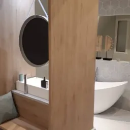 Nowoczesna łazienka z drewnianą zabudową umywalki, okrągłym lustrem na zawieszeniu i wolnostojącą wanną na tle wzorzystej ściany.