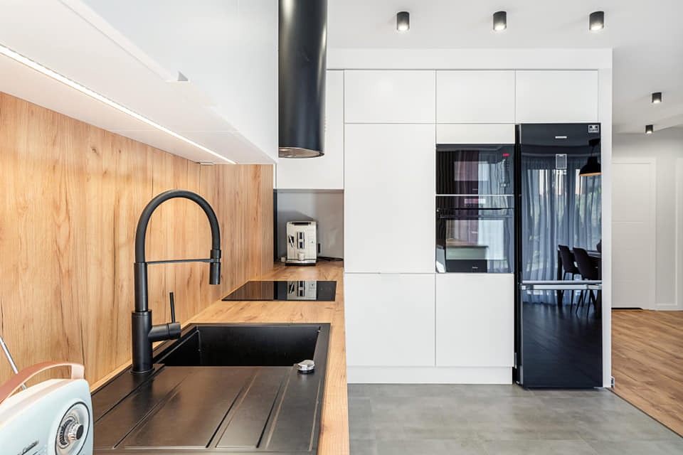Nowoczesna kuchnia z drewnianą okładziną ścienną, czarnym zlewem i baterią, oraz białymi frontami szafek z wbudowanym piekarnikiem i czarną lodówką.