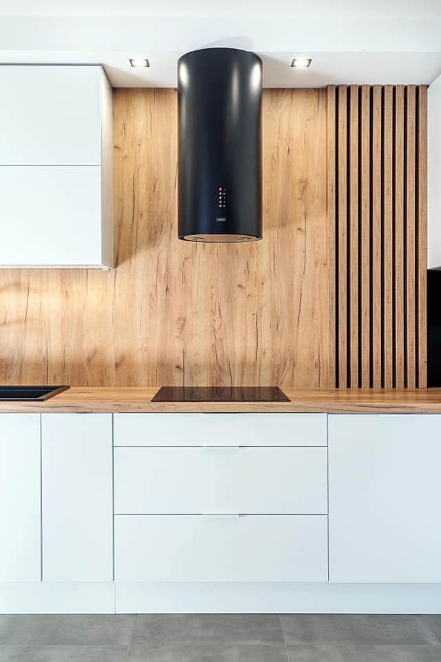 Nowoczesna kuchnia z białymi frontami, drewnianym blatem i panelem ściennym imitującym drewno, uzupełniona czarnym, cylindrycznym okapem.