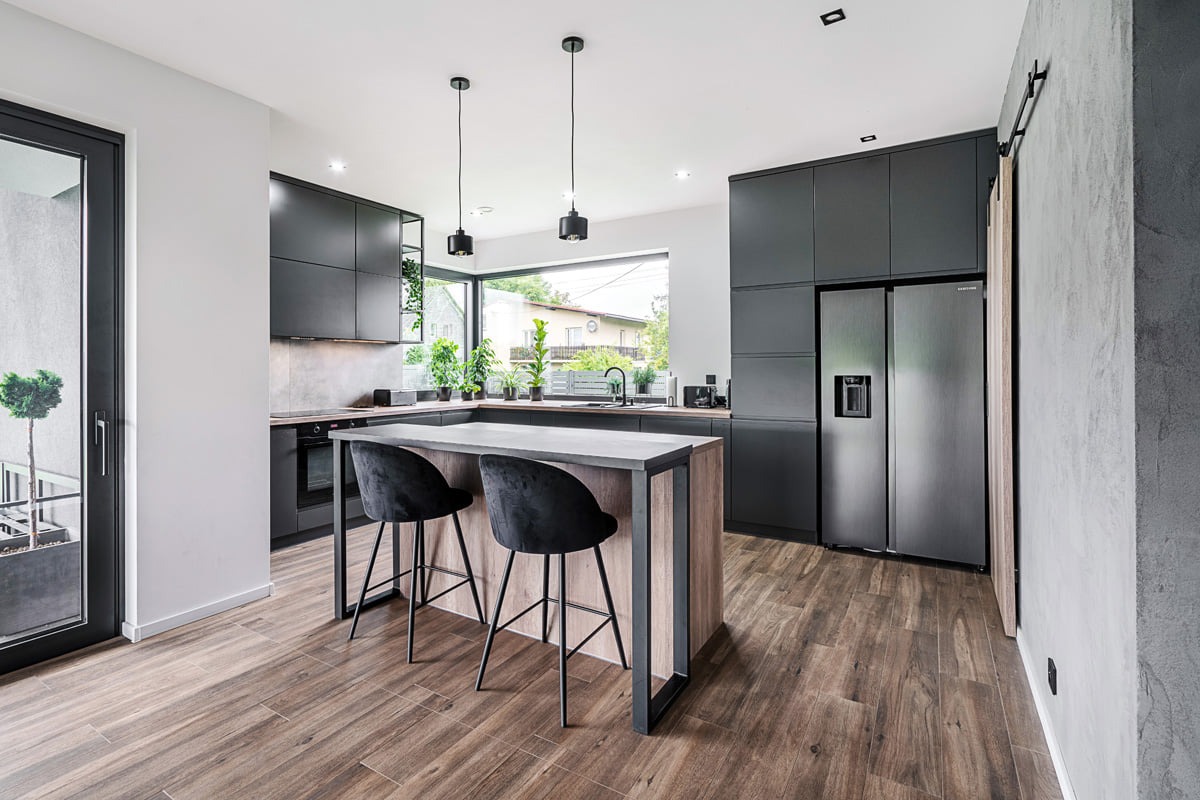 Nowoczesna kuchnia z czarnymi matowymi szafkami, wyspą z drewnianym wykończeniem i czarnymi hokerami, oświetlona wiszącymi lampami, widok na balkon i ogród przez duże okno.