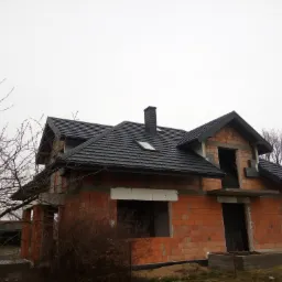 Niedokończony dom murowany z czarnym dachem i kominem, widok z zewnątrz w pochmurny dzień, drzewa bez liści w tle.