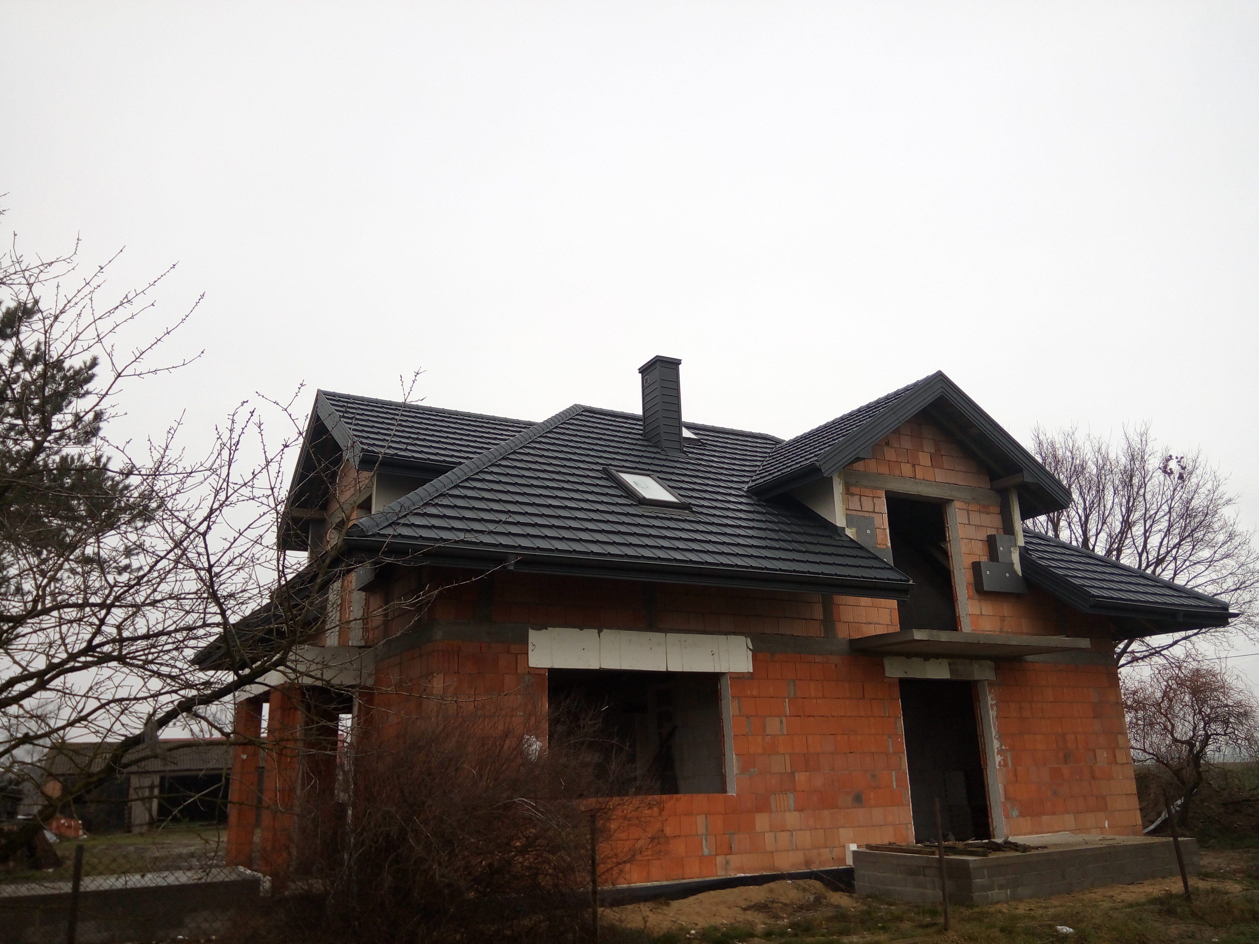 Niedokończony dom murowany z czarnym dachem i kominem, widok z zewnątrz w pochmurny dzień, drzewa bez liści w tle.