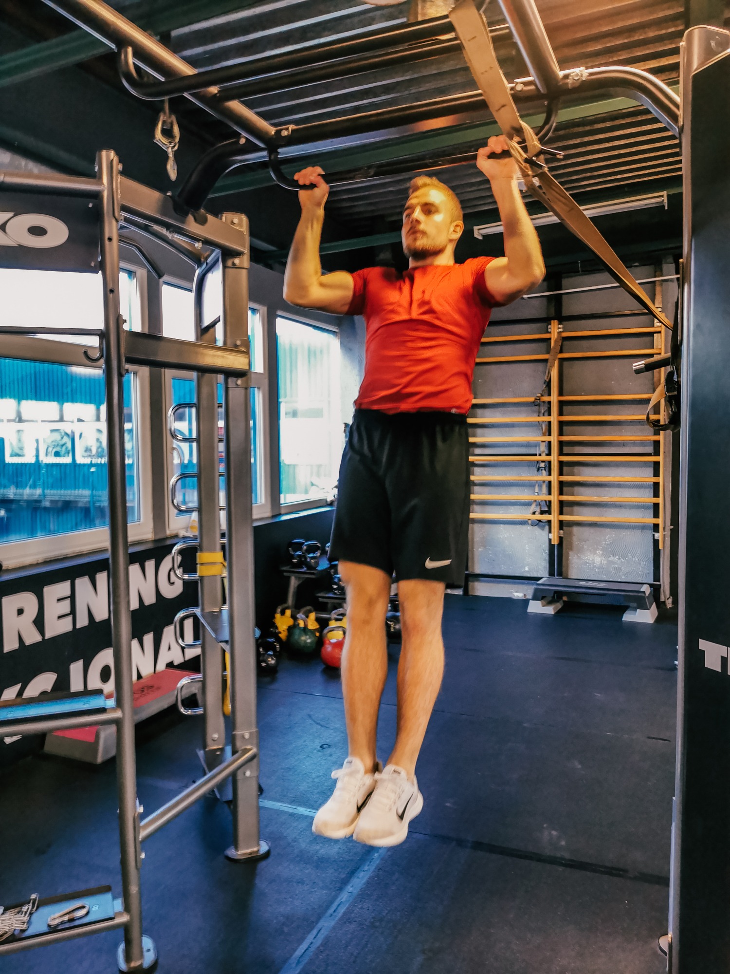 Mężczyzna w czerwonej koszulce i czarnych spodenkach wykonuje podciąganie na drążku w siłowni, widoczne drabinki gimnastyczne i kettlebells w tle.