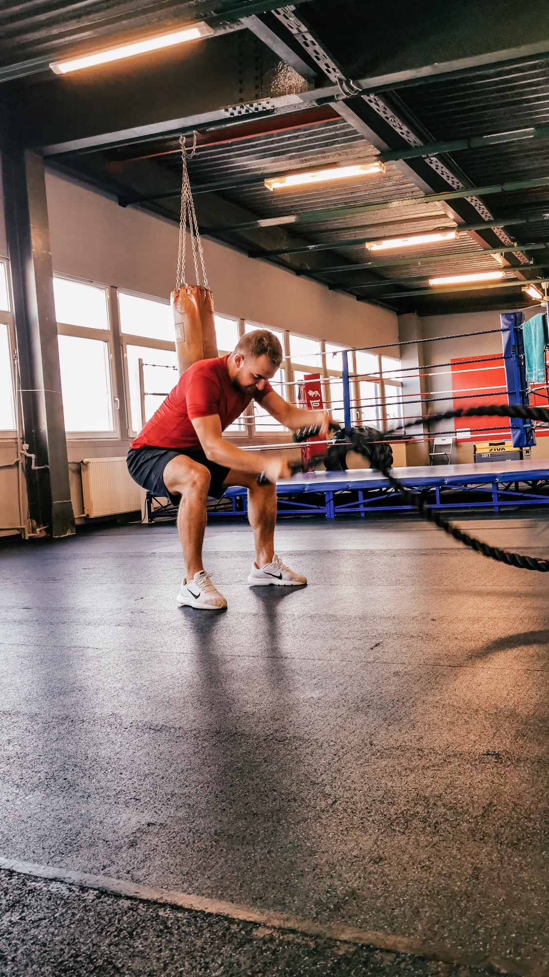 Mężczyzna w czerwonej koszulce ćwiczy z linami w sali treningowej z workiem bokserskim w tle.
