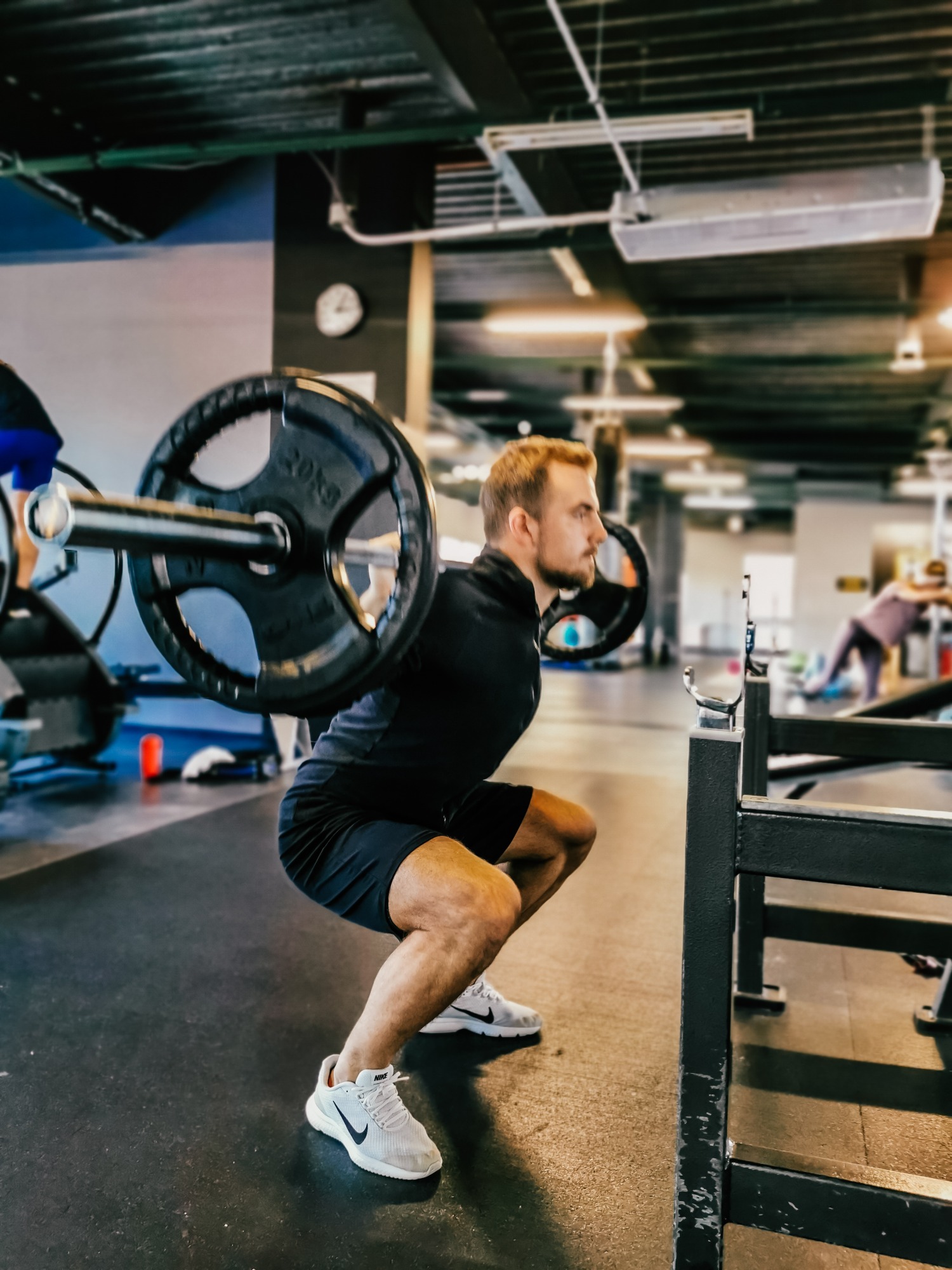 Mężczyzna w czarnym stroju wykonuje przysiad ze sztangą w siłowni, widoczne obciążenia 20kg, w tle inne osoby ćwiczące.