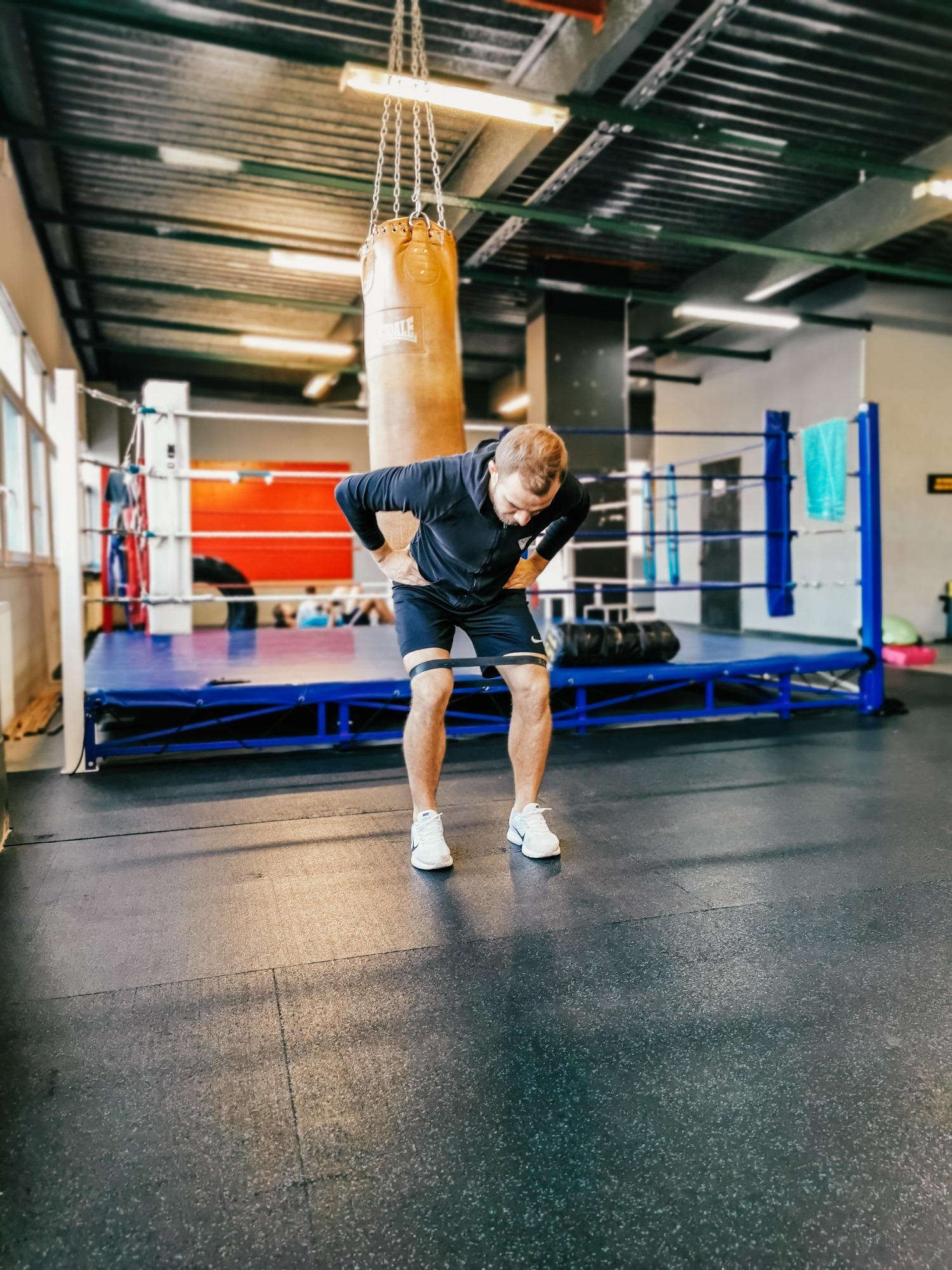 Mężczyzna w sportowym stroju wykonuje ćwiczenie z gumą oporową na nogach w pobliżu ringu bokserskiego i worka treningowego.