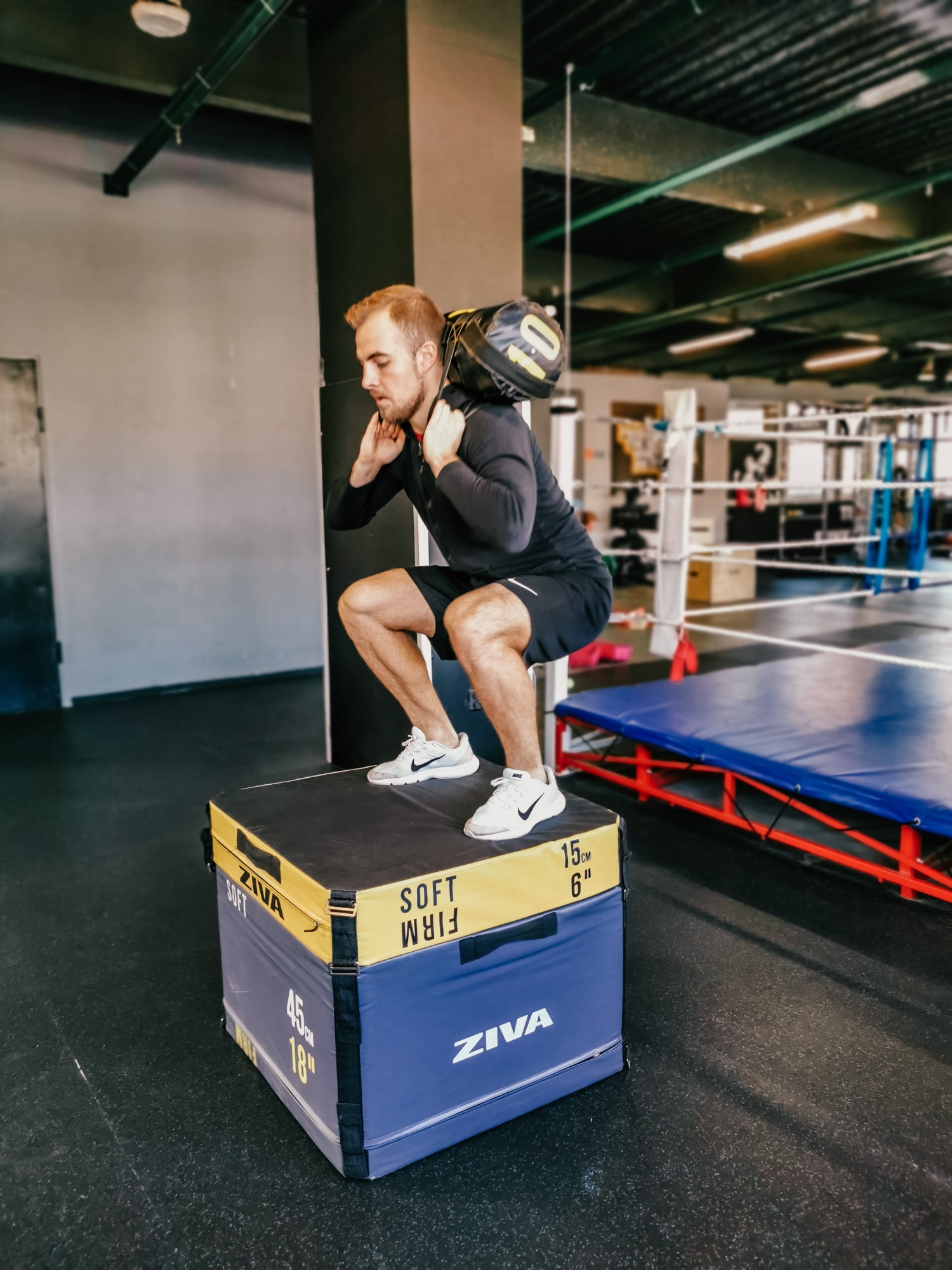 Mężczyzna w czarnym stroju sportowym wykonuje przysiad na skrzyni Ziva z obciążeniem na ramionach w sali gimnastycznej.