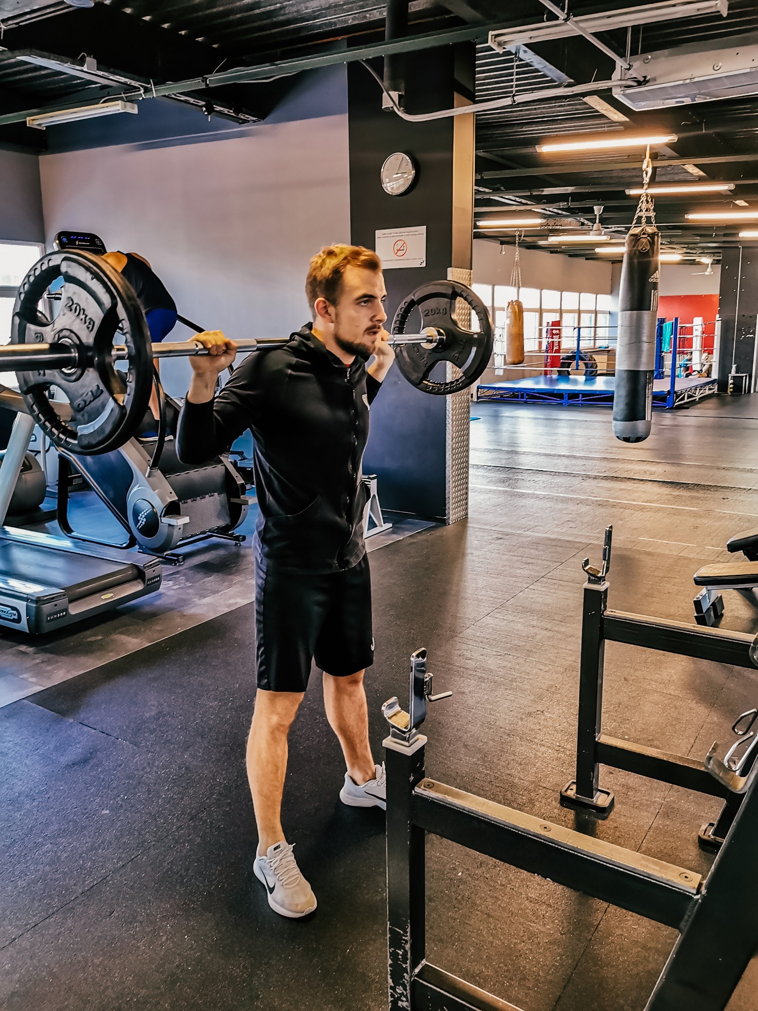 Mężczyzna w czarnym dresie wykonuje przysiad ze sztangą w siłowni z workami bokserskimi i bieżnią w tle.