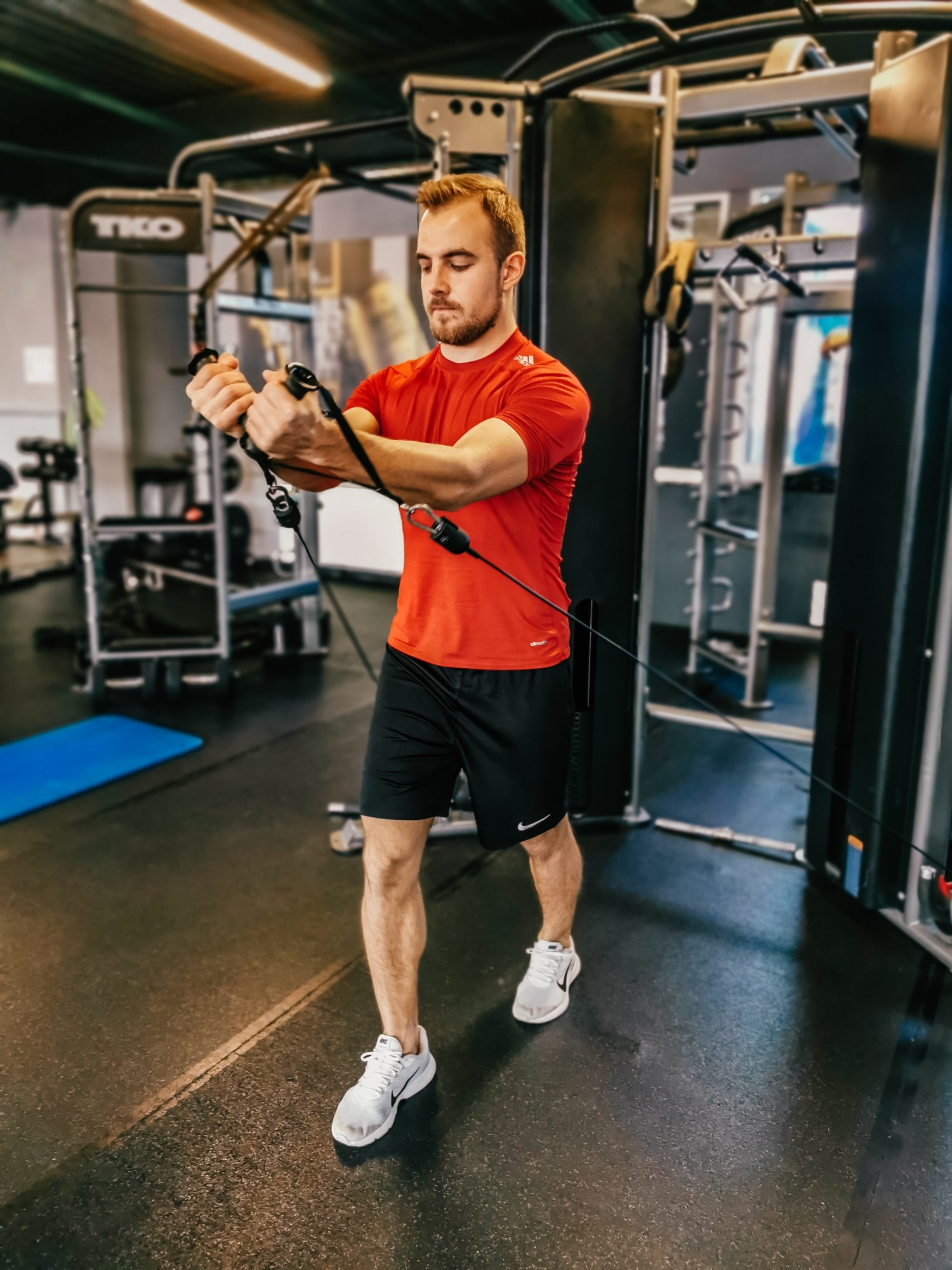 Mężczyzna w czerwonej koszulce ćwiczy na maszynie wyciągowej w siłowni, skupiony wzrok, białe buty sportowe.