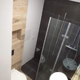 Nowoczesna łazienka z kabiną prysznicową ze szklanymi drzwiami, brązowymi płytkami i drewnopodobną ścianą z wnęką.