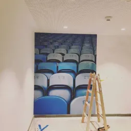 Duża fototapeta z motywem trybun stadionu na ścianie biura, z drewnianą drabiną opartą o ścianę i perforowanym sufitem podwieszanym.