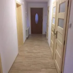 Wnętrze korytarza z nową podłogą z płytek imitujących drewno, świeżo pomalowanymi ścianami w odcieniach bieli i beżu, oraz surowymi drewnianymi drzwiami z przeszkleniami.