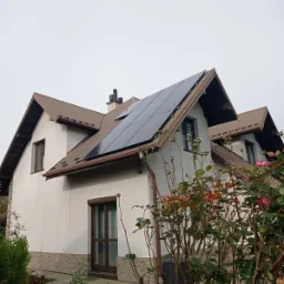 Panele słoneczne na brązowym dachu dwuspadowym domu jednorodzinnego z beżową elewacją, widoczne z poziomu ogrodu z krzewami róż.