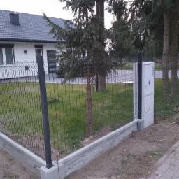 Nowoczesne ogrodzenie panelowe z podmurówką betonową i słupkami w kolorze antracytowym, widoczne skrzynki elektryczne na słupku, w tle dom jednorodzinny i iglaki.