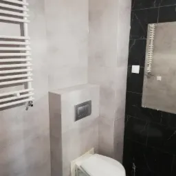 Nowoczesna łazienka z szarymi płytkami imitującymi beton, czarnymi płytkami z białymi żyłami, podwieszaną toaletą z chromowanym przyciskiem spłukiwania i białym grzejnikiem drabinkowym.