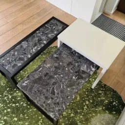 Zestaw trzech stolików kawowych: prostokątny z czarną ramą i blatem imitującym szary marmur, kwadratowy z blatem imitującym szary marmur oraz biały kwadratowy stolik, ustawione na zielonym dywanie...