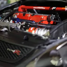 Otwarta maska sportowego samochodu Honda Civic Type R z widocznym, zmodyfikowanym silnikiem: czerwona pokrywa, aluminiowa rura dolotowa owinięta folią termoizolacyjną, czerwona rozpórka kielichów...