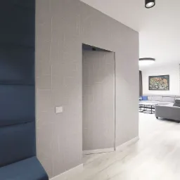 Nowoczesny hol apartamentu z ukrytymi drzwiami w ścianie pokrytej geometryczną tapetą, obok niebieski panel ścienny, widok na salon z szarą sofą i obrazem.