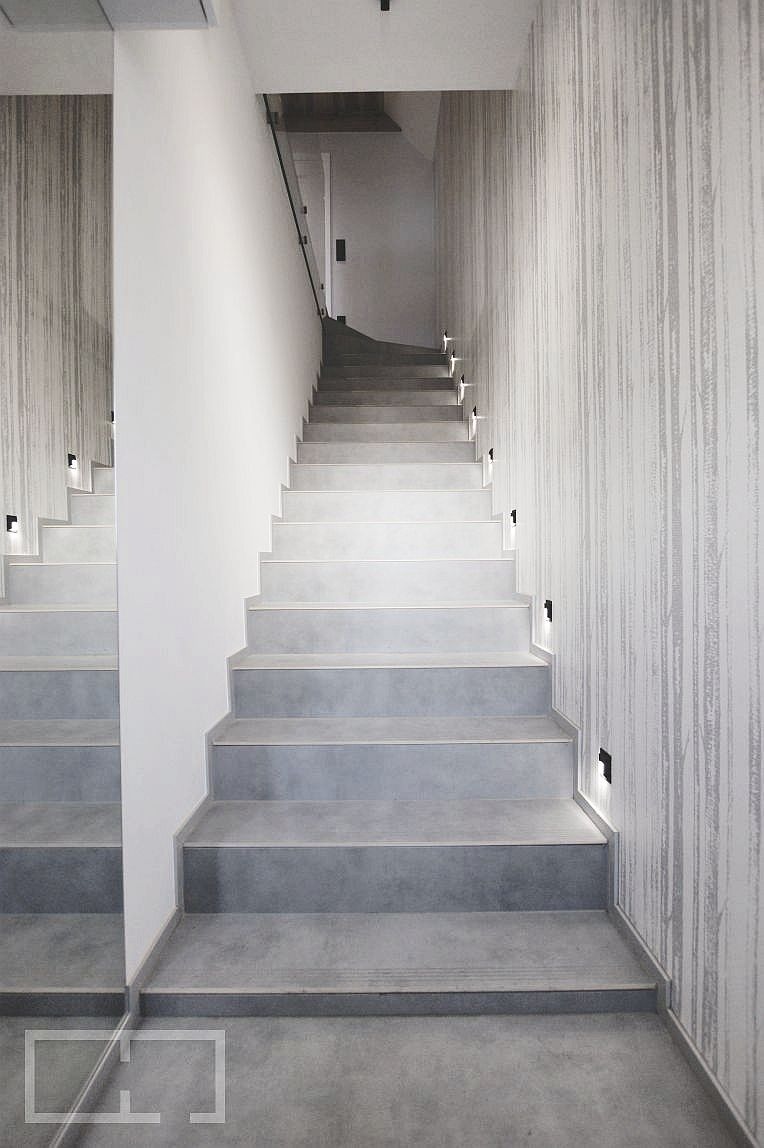 Nowoczesne schody wewnętrzne z szarymi stopniami i minimalistycznym oświetleniem punktowym, ściana pokryta tapetą w subtelny wzór drzew.