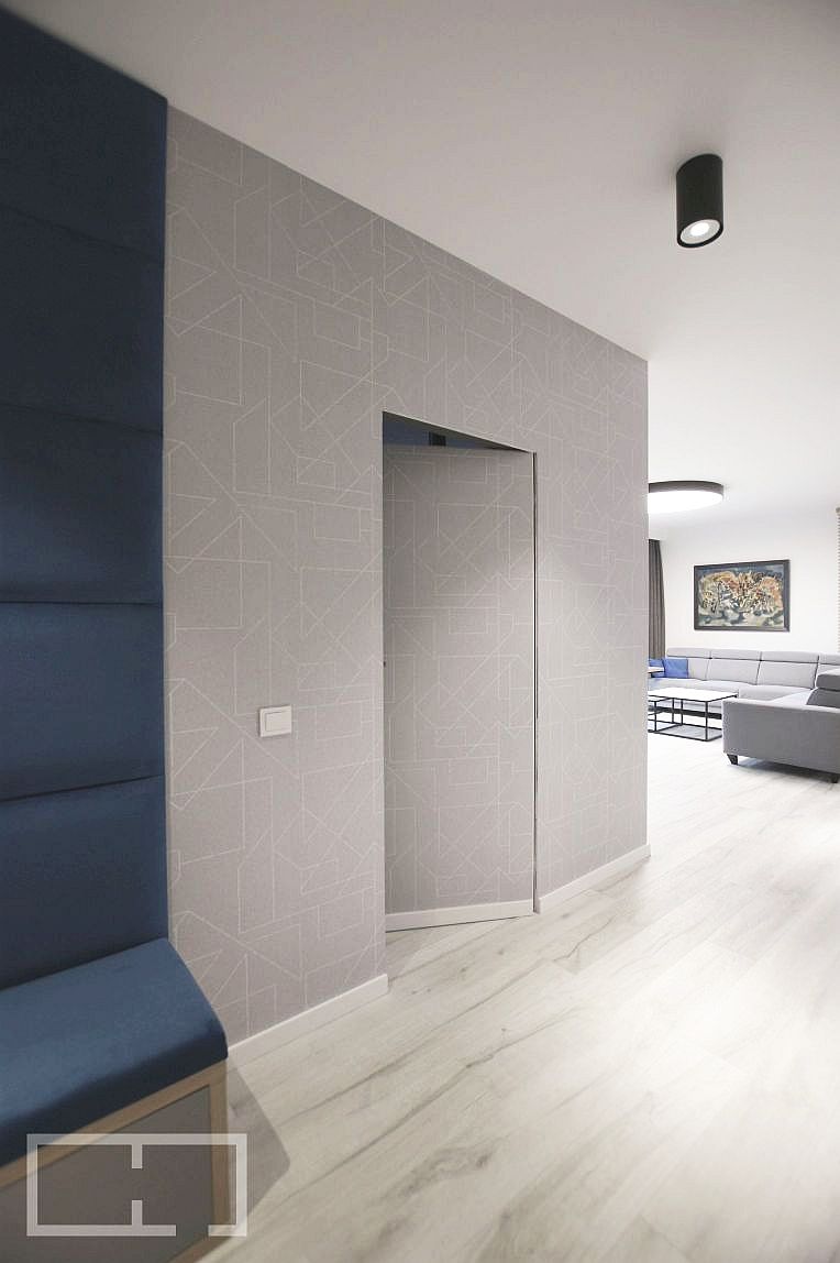 Nowoczesny hol apartamentu z ukrytymi drzwiami w ścianie pokrytej geometryczną tapetą, obok niebieski panel ścienny, widok na salon z szarą sofą i obrazem.
