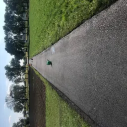 Nowo wyasfaltowana droga biegnąca przez teren zielony, widoczne drzewa i niebieskie niebo z lekkimi chmurami, zielony kanister na drodze.