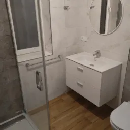 Nowoczesna łazienka z kabiną prysznicową, białą umywalką z szafką, okrągłym lustrem i drewnopodobną podłogą, wykończona płytkami imitującymi marmur.