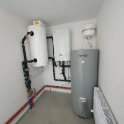 Biały zasobnik ciepłej wody z czarnymi rurami i zaworami, obok biały piec gazowy LG Therma V, szary zasobnik Ferroli i biały grzejnik panelowy na tle białych ścian i szarej podłogi.