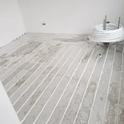 Białe rurki ogrzewania podłogowego ułożone na szarym podłożu w pustym, jasnym pomieszczeniu. Obok zwój rurek. Widoczne ślady po montażu.