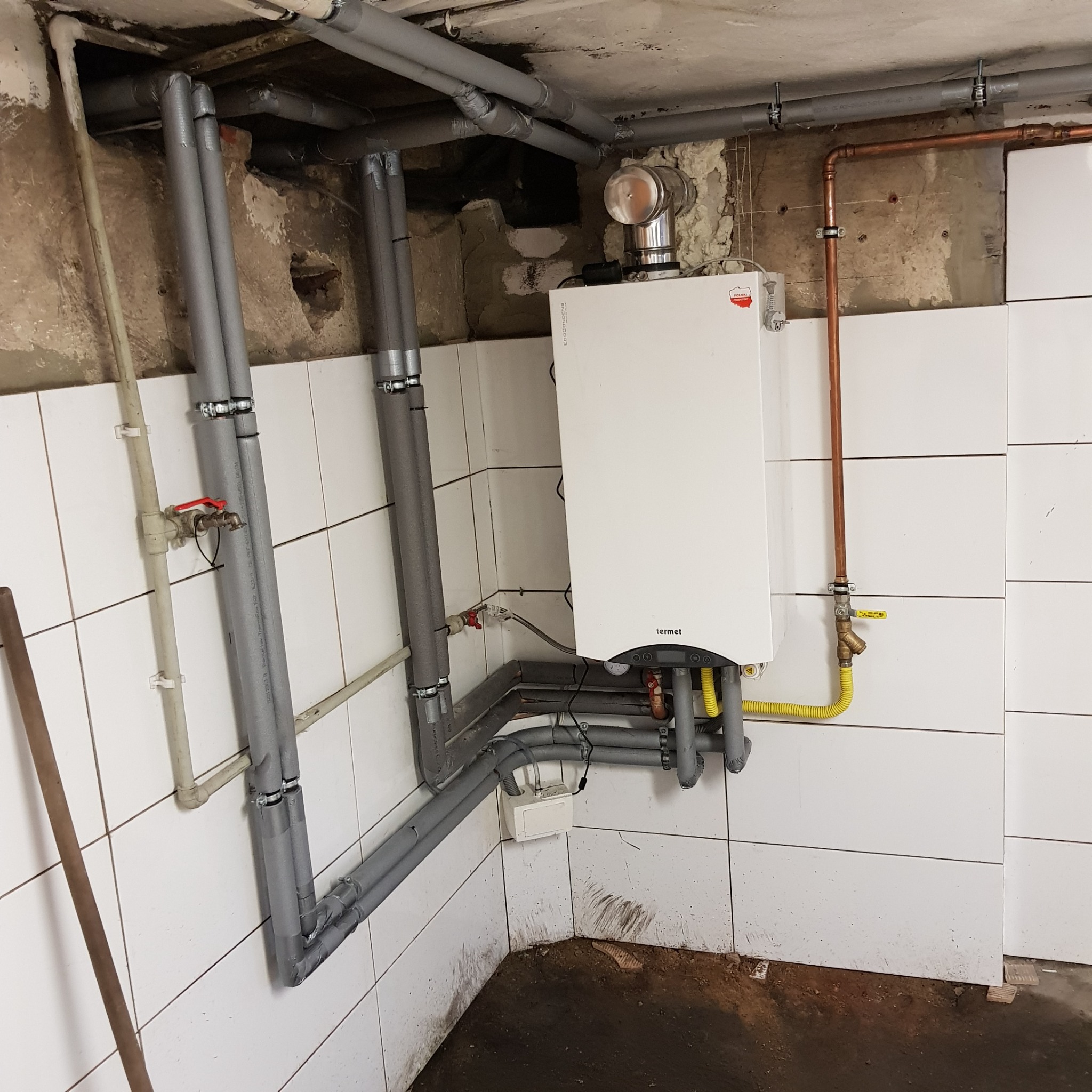 Biały piec gazowy Termet z podłączeniami, rurami izolowanymi i instalacją miedzianą na tle białych płytek w pomieszczeniu w trakcie prac instalacyjnych.