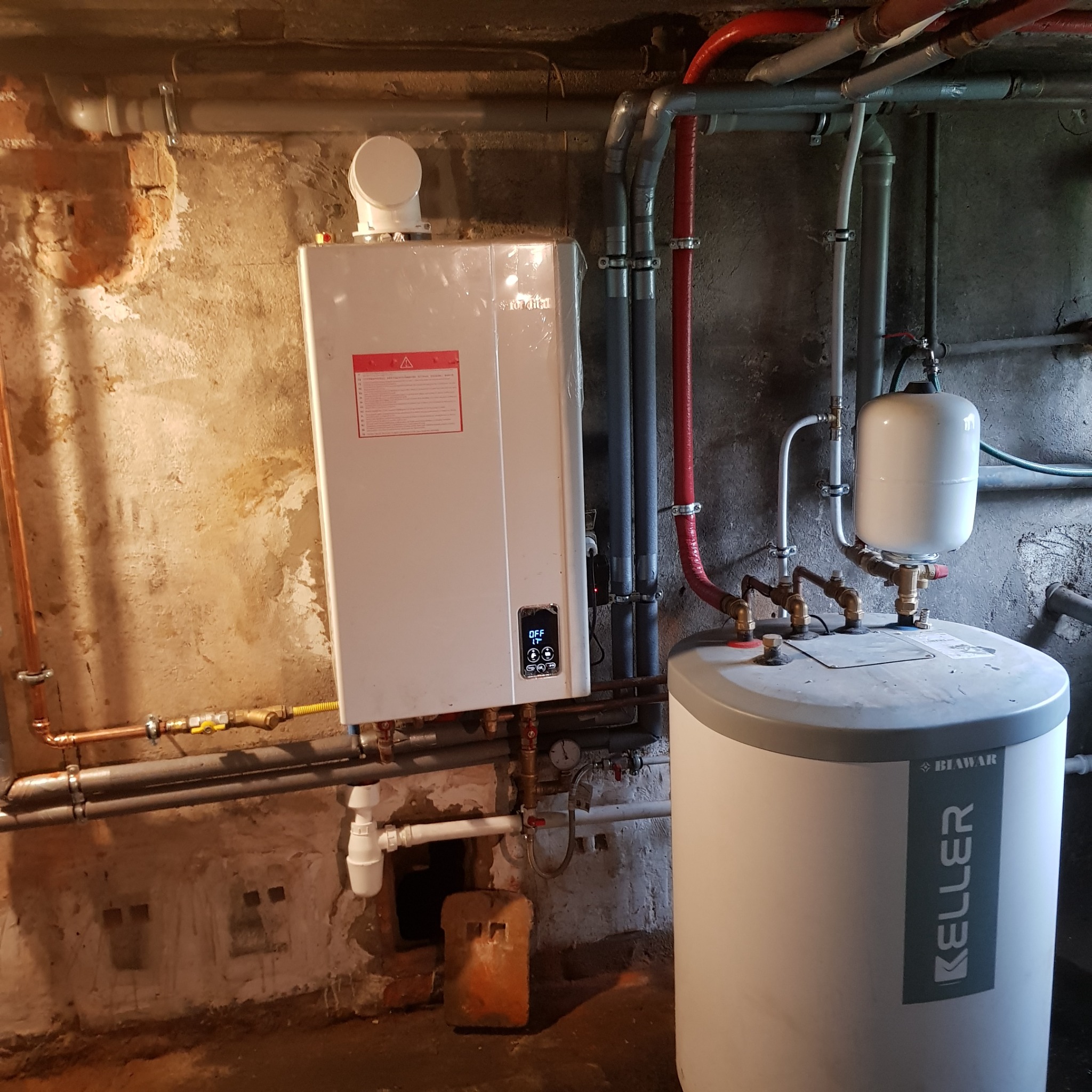 Nowoczesny piec gazowy i zasobnik ciepłej wody marki Biawar Keller, zamontowane w piwnicy z widocznymi rurami miedzianymi i plastikowymi.