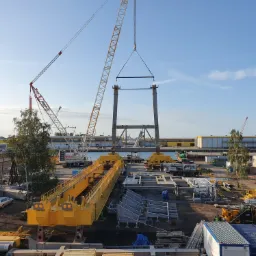 Montaż stalowej konstrukcji rozładowarki statków przy użyciu dźwigów na nabrzeżu portowym, widoczne elementy konstrukcyjne w kolorze żółtym i szarym, oraz mniejsze maszyny budowlane.