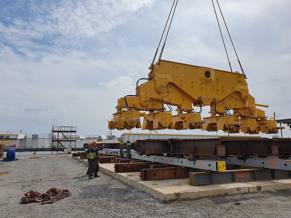 Żółty element rozładowarki statków podnoszony przez linę dźwigu nad stalowymi belkami, dwóch pracowników w kaskach i kamizelkach obserwuje proces na placu budowy.