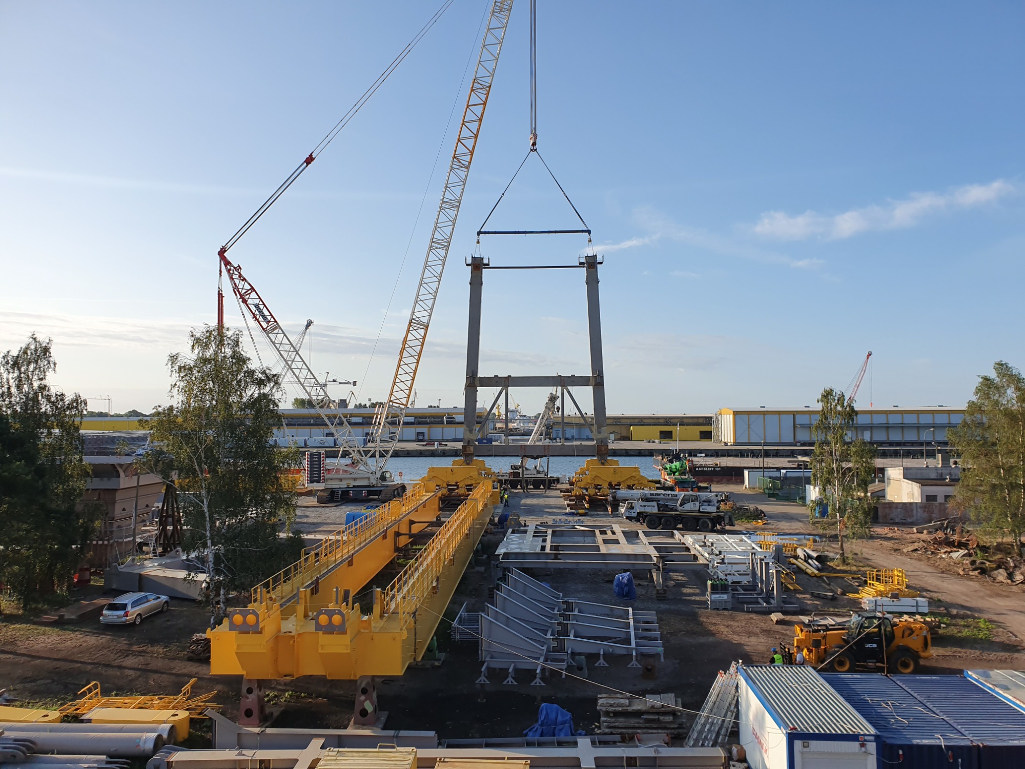 Montaż stalowej konstrukcji rozładowarki statków przy użyciu dźwigów na nabrzeżu portowym, widoczne elementy konstrukcyjne w kolorze żółtym i szarym, oraz mniejsze maszyny budowlane.