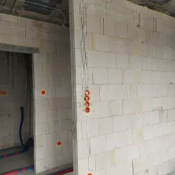 Surowe ściany z betonu komórkowego w trakcie budowy, widoczne białe przewody elektryczne ułożone w korytach, pomarańczowe puszki instalacyjne, oraz rury instalacji wodnej w kolorze niebieskim...