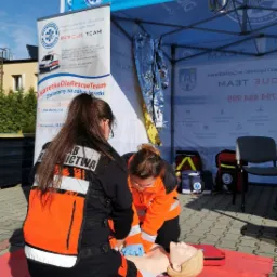 Instruktorzy w odzieży roboczej demonstrują resuscytację krążeniowo-oddechową na fantomie podczas plenerowego pokazu, widoczne logo Rescue Team na banerze w tle.