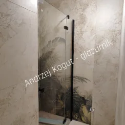 Kabina prysznicowa z czarnymi okuciami i wzorem liści na płytkach w beżowej łazience. Widoczne przyłącza wody i fragment butelki na podłodze.