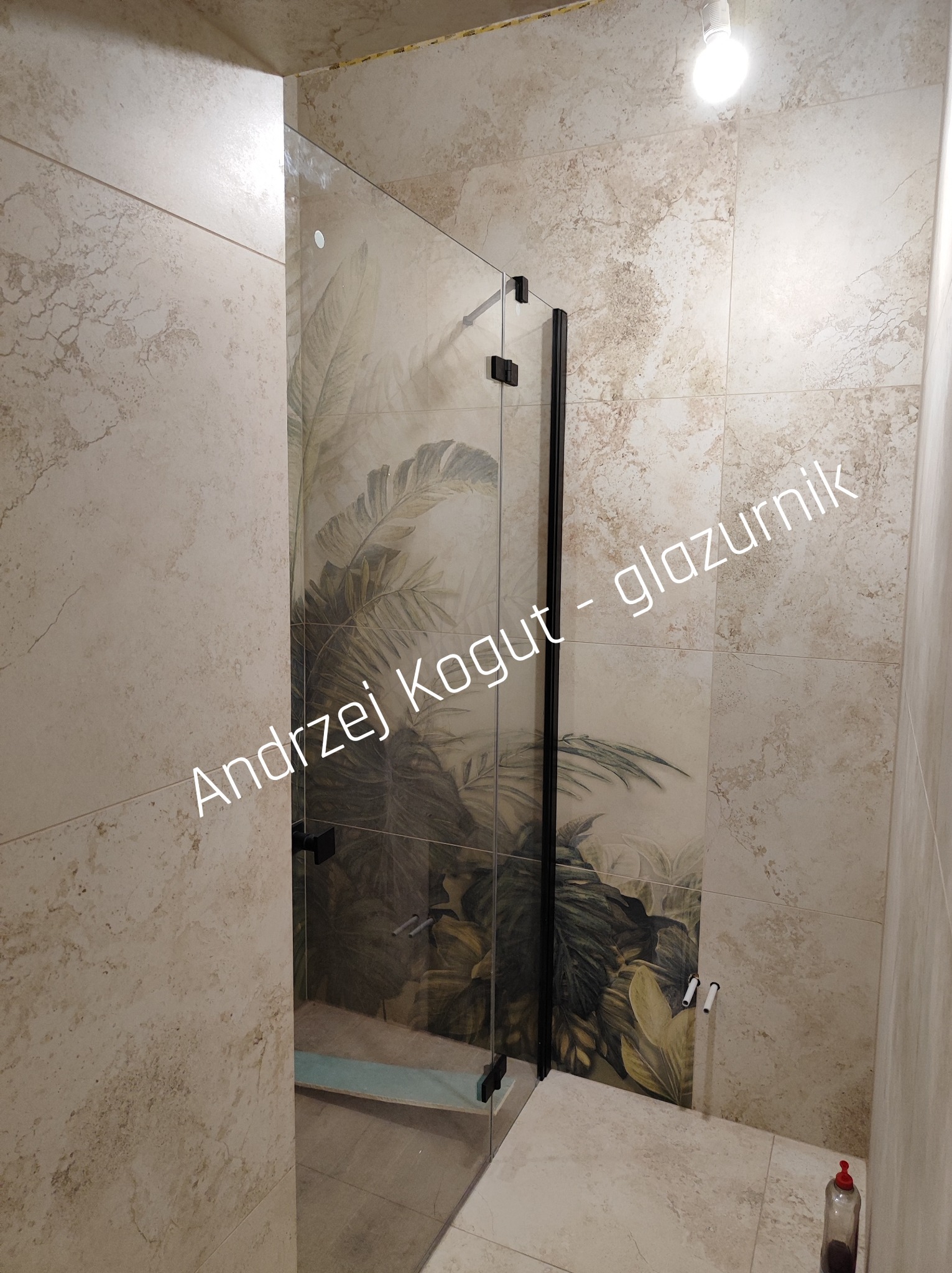 Kabina prysznicowa z czarnymi okuciami i wzorem liści na płytkach w beżowej łazience. Widoczne przyłącza wody i fragment butelki na podłodze.