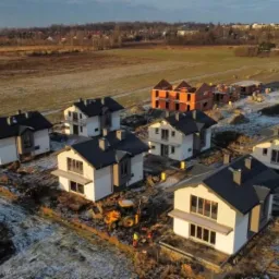 Osiedle domów jednorodzinnych w trakcie budowy, widok z lotu ptaka, częściowo wykończone elewacje, teren przygotowany pod zagospodarowanie, w tle krajobraz podmiejski.