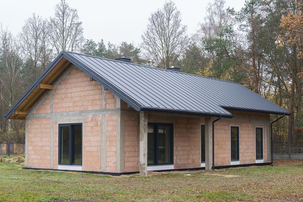 Niedokończony parterowy dom z czerwonej cegły z szarą metalową blachodachówką, widoczne ocieplenie fundamentów i wstępnie osadzone okna, na tle jesiennego krajobrazu.