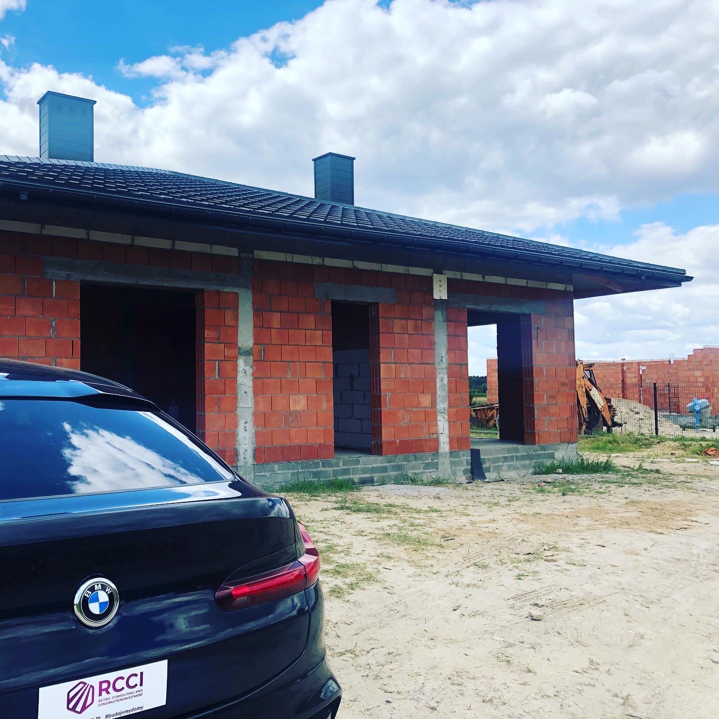 Budowa domu w trakcie wznoszenia ścian z czerwonej cegły, widoczny fragment dachu z kominami oraz samochód BMW z logo firmy budowlanej w tle.