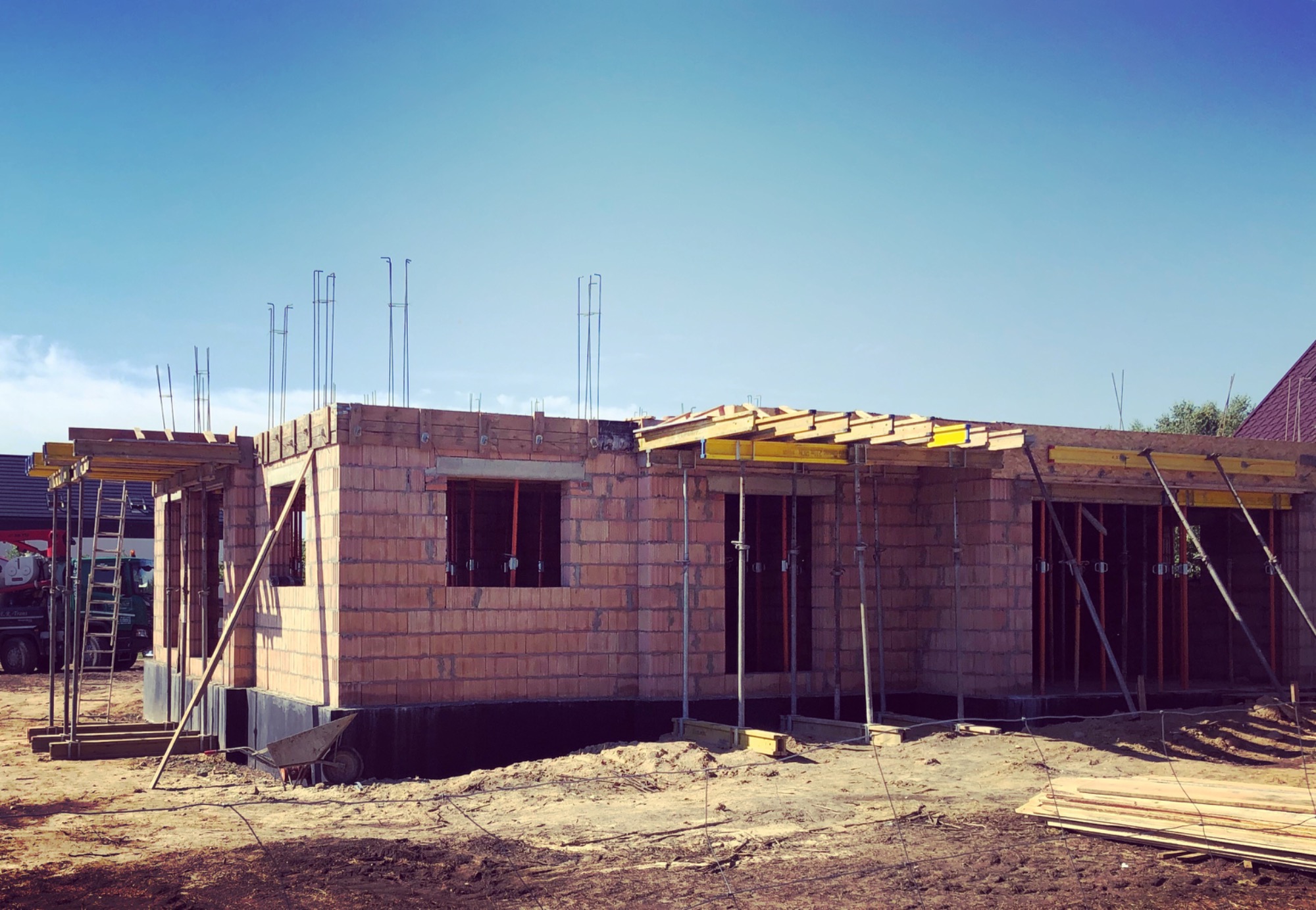 Stan surowy otwarty domu jednorodzinnego w Łodzi, z widocznymi elementami szalunków i zbrojenia, wykonany z czerwonej cegły. Na drugim planie widoczny samochód ciężarowy i drabina.