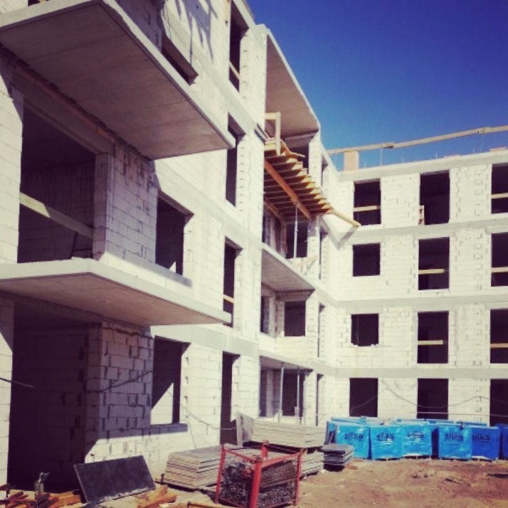 Wielopiętrowy budynek w trakcie wznoszenia z białych bloków, widoczne niezamurowane otwory okienne i balkony, na dole palety z materiałami budowlanymi.