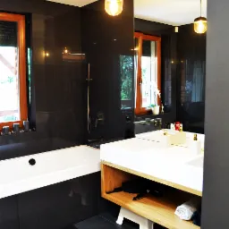 Nowoczesna łazienka z czarnymi płytkami, drewnianą ramą okienną i białym blatem umywalki z podświetleniem, widok na wannę i szafkę pod umywalką.