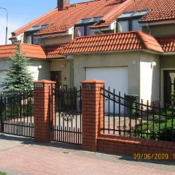 Domy szeregowe z czerwoną dachówką, czarnym ogrodzeniem z ceglanymi słupkami i zielonymi elewacjami w słoneczny dzień.