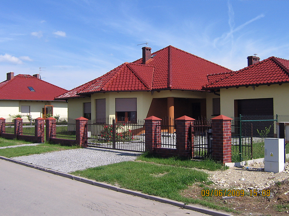 Nowoczesny dom jednorodzinny z czerwoną dachówką, ogrodzony metalowym płotem z ceglanymi słupkami, widok z ulicy w słoneczny dzień.