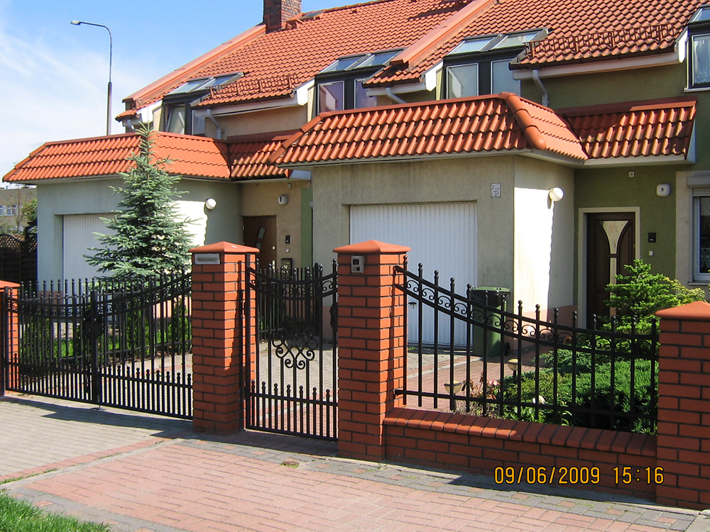 Domy szeregowe z czerwoną dachówką, czarnym ogrodzeniem z ceglanymi słupkami i zielonymi elewacjami w słoneczny dzień.