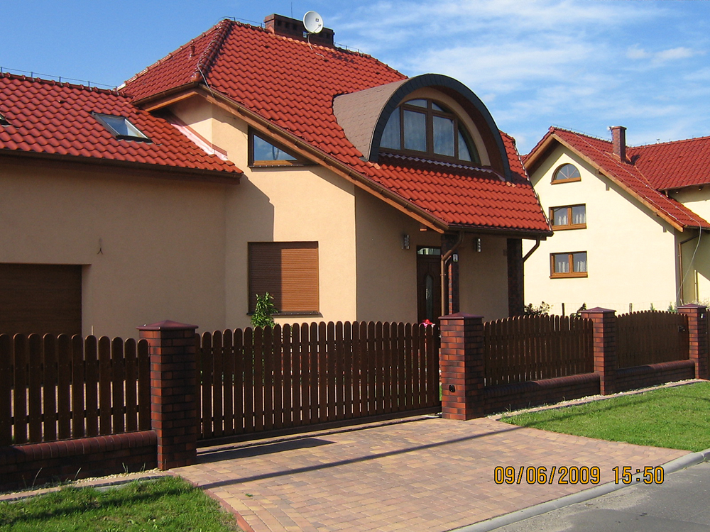 Nowoczesny dom jednorodzinny z czerwoną dachówką, drewnianym ogrodzeniem z ceglanymi słupkami i brukowanym podjazdem w słoneczny dzień.